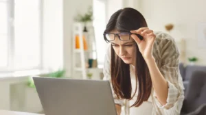 Woman lifting her glasses while looking closely at a laptop screen, suggesting difficulty reading or comparing vision correction options.